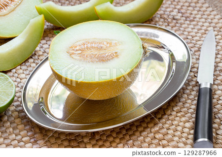 Diagonally at the center on a silver platter half of melon near melon slices, knife on a straw napkin background. Half of melon on silver platter. Horizontal. 129287966