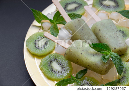 Chilled kiwi icecream popsicles are presented on a golden plate surrounded by ice cubes and fresh kiwi slices, creating a refreshing summer delight at a gathering. Chilled kiwi icecream popsicles are presented on a golden plate surrounded by ice cubes and fresh kiwi slices, creating a refreshing summer delight at a gathering. 129287967