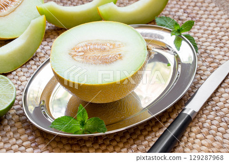 Diagonally at the center on a silver platter half of melon near melon slices, mint leaves, knife on a straw napkin background. Half of melon on silver platter. Horizontal. Close. 129287968