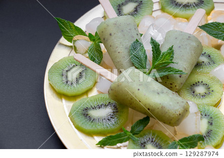 Three refreshing kiwi ice cream pops are elegantly displayed on a golden plate surrounded by sliced kiwis and garnished with fresh mint leaves, creating a vibrant summer treat. Three refreshing kiwi ice cream pops are elegantly displayed on a golden plate surrounded by sliced kiwis and garnished with fresh mint leaves, creating a vibrant summer treat. 129287974