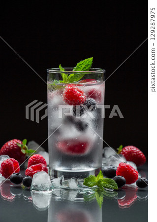 A glass of chilled mineral water with ice cubes and berries near spilled blueberries, strawberries, raspberries and ice cubes on a black background. Mineral water with berries and ice cubes. Vertical. A glass of chilled mineral water with ice cubes and berries near spilled blueberries, strawberries, raspberries and ice cubes on a black background. Mineral water with berries and ice cubes. Vertical. 129287975