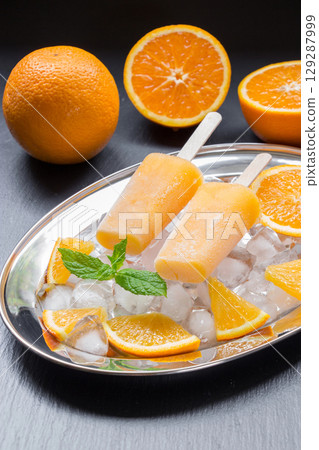 Orange sorbet ice cream on silver plate with ice cubes, orange pieces, mint leaves near orange slices on a black background. Orange fruit sorbet ice cream popsicles. Vertical. 129287999