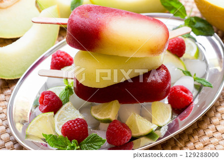 Vibrant melon raspberry ice cream sits atop a mound of ice cubes, surrounded by fresh raspberries, mint leaves, and sliced lime. 129288000