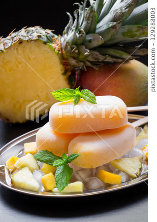 Exotic sorbet ice cream popsicles on silver plate with ice cubes chopped pineapple mango pieces, mint near half of pineapple, mango on a black background. Pineapple mango sorbet ice cream popsicles. 129288003