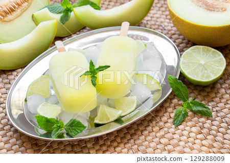 Enjoy refreshing melon ice cream popsicles presented on a silver tray surrounded by ice, mint, and lime, ideal for a warm afternoon gathering with friends or family. Enjoy refreshing melon ice cream popsicles presented on a silver tray surrounded by ice, mint, and lime, ideal for a warm afternoon gathering with friends or family. 129288009