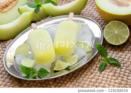 Melon and lime sorbet ice cream on silver plate with ice cubes, lime pieces, mint leaves near melon slices, lime on a straw napkin background. Melon and lime sorbet ice cream popsicles. Horizontal. 129288016