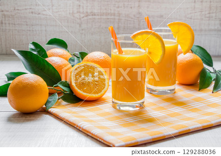 Two tall glasses of orange juice are placed next to fresh oranges on a checkered cloth. The bright kitchen setting is enhanced by natural daylight, creating a cheerful atmosphere. 129288036