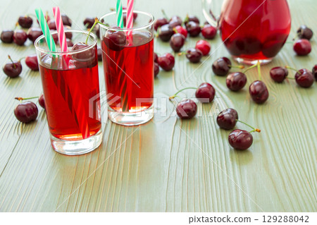 Left 2 glasses of juice, green straws, scattered cherries, jar of cherry juice, on right empty space for text on green background. Cherries, cherry juice and empty space. Horizontal. 129288042