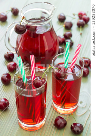Two glasses of cherry juice with straws in the foreground and in the background a jar full of cherry juice. 2 cherry juices. Vertical. Close-up. Daylight. Two glasses of cherry juice with straws in the foreground and in the background a jar full of cherry juice. 2 cherry juices. Vertical. Close-up. Daylight. 129288070