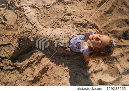 Child enjoys a sunny day at the beach, creating a sand mermaid sculpture 129288263
