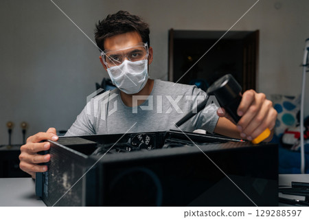 Portrait of technician in face mask and safety glasses looking at camera cleaning computer case using small vacuum cleaner, demonstrating proper maintenance procedures. Concept of service computer. 129288597