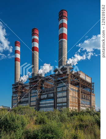 A large power plant with four red and white chimney towers stands against the blue sky, surrounded by lush green vegetation. 129289162