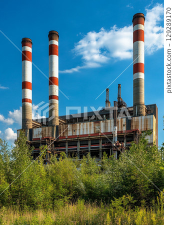 A large power plant with four red and white chimney towers stands against the blue sky, surrounded by lush green vegetation. 129289170