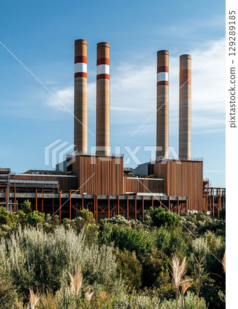 A large power plant with four red and white chimney towers stands against the blue sky, surrounded by lush green vegetation. 129289185
