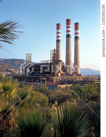 A large power plant with four red and white chimney towers stands against the blue sky, surrounded by lush green vegetation. 129289186
