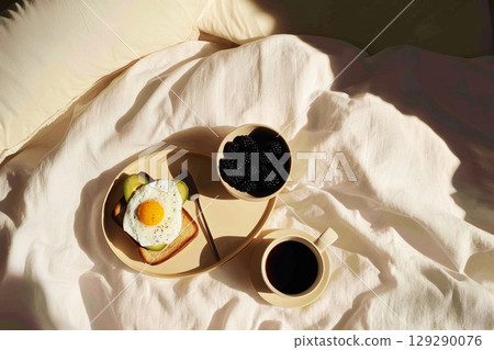 Soft morning light streams in as a delicious breakfast spread is laid out on a cozy bed. A plate features toasted bread topped with an egg, with berries and coffee on the side 129290076