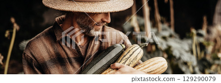 A farmer in a straw hat gathers an assortment of squash from the autumn garden. The warm, earthy colors of the season create a serene backdrop for this harvest activity A farmer in a straw hat gathers an assortment of squash from the autumn garden. The warm, earthy colors of the season create a serene backdrop for this harvest activity 129290079