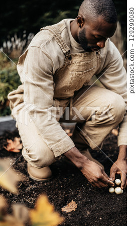 A person kneels on the earth, carefully placing mushroom spores in neat rows. Nearby, vibrant daffodils sway gently in the autumn breeze, surrounded by fallen leaves 129290080