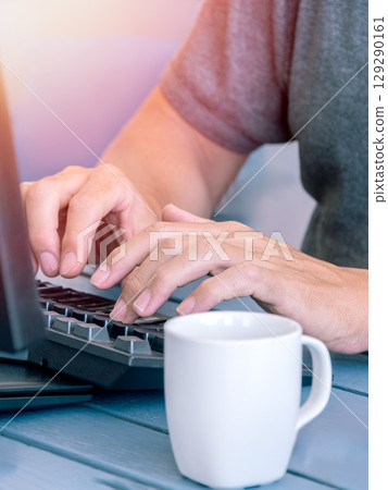 Hands typing on keyboard with coffee cup nearby 2.- Close-up of hands typing on a keyboard with a white coffee cup on a blue table. Hands typing on keyboard with coffee cup nearby 2.- Close-up of hands typing on a keyboard with a white coffee cup on a blue table. 129290161