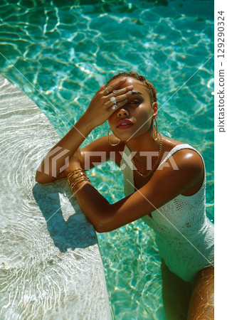 A woman in a white bikini is sitting on a poolside ledge, looking at the camera 129290324