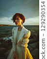 A woman in a white dress stands on a rocky shoreline 129290354