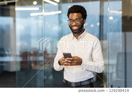 Professional man wearing glasses holds a smartphone while smiling in a glass-walled office environment, capturing a moment of productivity and confidence. Professional man wearing glasses holds a smartphone while smiling in a glass-walled office environment, capturing a moment of productivity and confidence. 129290419