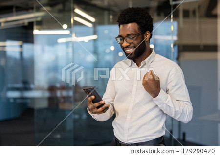 A smiling professional man in a modern office celebrates while holding his phone. The image represents achievement, inspiration, and positive energy in a workplace environment. A smiling professional man in a modern office celebrates while holding his phone. The image represents achievement, inspiration, and positive energy in a workplace environment. 129290420