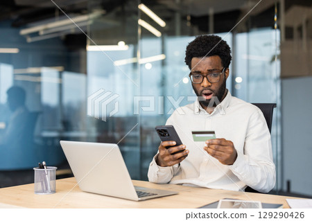 Professional man at a desk in a modern office, surprised by credit card details on his phone. Professional man at a desk in a modern office, surprised by credit card details on his phone. 129290426