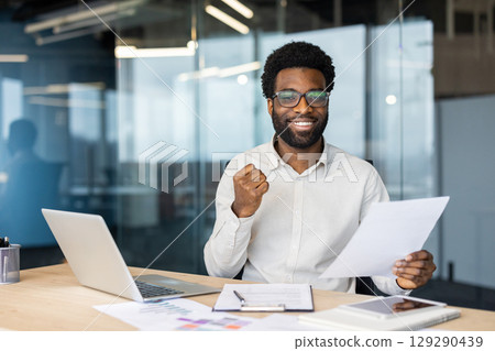 Professional man expressing confidence and success while working at his desk in an office. He holds documents and uses a laptop, showcasing a productive and motivated work attitude. 129290439