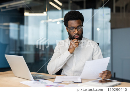 A businessman attentively examines paperwork at his desk in a contemporary office. He is surrounded by modern tools, including a laptop and smartphone, highlighting a setting of productivity 129290440