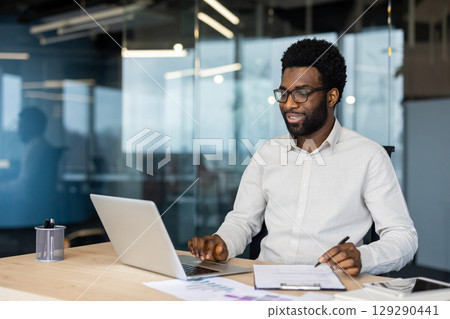 A focused individual working on a laptop at a desk in a contemporary office. The environment suggests productivity, professionalism, and a modern work setting with a hint of collaboration. A focused individual working on a laptop at a desk in a contemporary office. The environment suggests productivity, professionalism, and a modern work setting with a hint of collaboration. 129290441