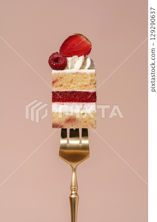 Vanilla sponge bento cake layered with raspberry and strawberry, topped with whipped cream and fresh fruit, presented on a golden fork against a soft pastel background, dessert concept 129290637