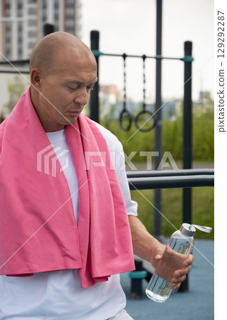 Serious sporty man tired and standing with water bottle at urban sport ground after workout. Summer outdoor air training. 129292287