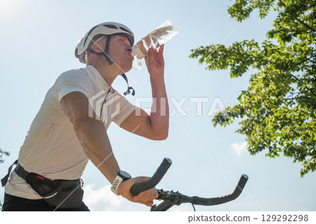 Professional male bike rider in white helmet drinks water against blue sky at city public park. Close up Professional male bike rider in white helmet drinks water against blue sky at city public park. Close up 129292298