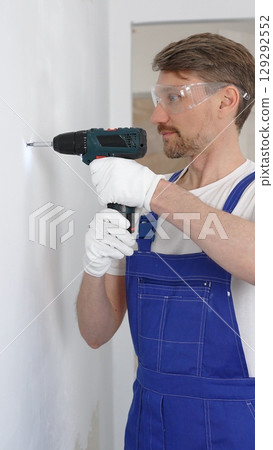 Professional male construction worker in a blue jumpsuit, safety goggles, and work gloves is screwing a screw into the wall using a cordless screwdriver, carrying out a careful home renovation Professional male construction worker in a blue jumpsuit, safety goggles, and work gloves is screwing a screw into the wall using a cordless screwdriver, carrying out a careful home renovation 129292552