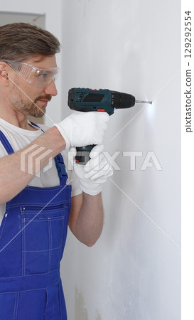 Professional male construction worker in a blue jumpsuit, safety goggles, and work gloves is screwing a screw into the wall using a cordless screwdriver, carrying out a careful home renovation Professional male construction worker in a blue jumpsuit, safety goggles, and work gloves is screwing a screw into the wall using a cordless screwdriver, carrying out a careful home renovation 129292554