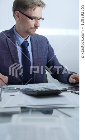 Businessman wearing blue suit is working with laptop, calculator and documents at the glass table in office set. Low lighting, vertical portrait. Business people and audit Businessman wearing blue suit is working with laptop, calculator and documents at the glass table in office set. Low lighting, vertical portrait. Business people and audit 129292555