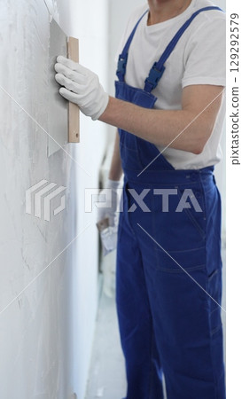 Man construction worker wearing protective gloves and blue construction coveralls, spreading plaster smoothly across wall using professional drywall taping knife Man construction worker wearing protective gloves and blue construction coveralls, spreading plaster smoothly across wall using professional drywall taping knife 129292579
