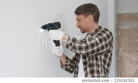 Man construction worker in checked shirt and safety gloves is screwing a screw into the wall using a cordless screwdriver, carrying out a careful home renovation. Horizontal portrait view 129292581