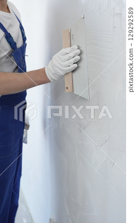 Male construction worker wearing protective gloves and blue construction coveralls, spreading plaster smoothly across wall using professional drywall taping knife during renovation, vertical view Male construction worker wearing protective gloves and blue construction coveralls, spreading plaster smoothly across wall using professional drywall taping knife during renovation, vertical view 129292589