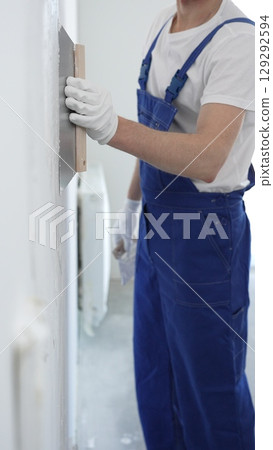Man construction worker wearing protective gloves and blue construction coveralls, spreading plaster smoothly across wall using professional drywall taping knife 129292594