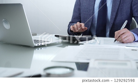 Businessman or accountant wearing blue suit is working with laptop, calculator and documents in office. Low lighting, close-up view. Business people, audit concept 129292598