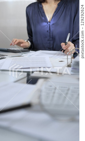 Businesswoman wearing blue dress is working with laptop, calculator and documents in a professional office setting. Low lighting, close-up vertical view. Business people, audit concept Businesswoman wearing blue dress is working with laptop, calculator and documents in a professional office setting. Low lighting, close-up vertical view. Business people, audit concept 129292624