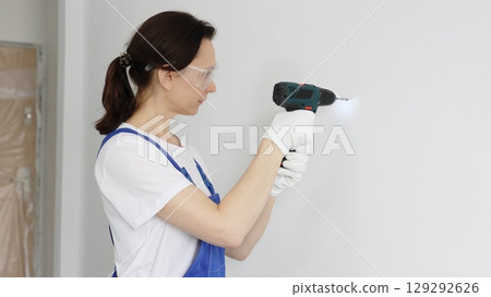 Professional female worker wearing blue overall, safety glasses and gloves, is using cordless drill to make hole in white wall during renovation work. Portrait view Professional female worker wearing blue overall, safety glasses and gloves, is using cordless drill to make hole in white wall during renovation work. Portrait view 129292626