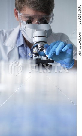 Man scientist researcher wearing blue gloves, protective glasses and medical mask is using microscope in modern laboratory, portrait view. Low key lighting photo. Science and Medicine concepts 129292630