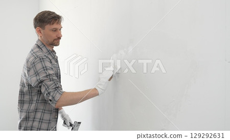 Construction worker wearing gloves and using a putty knife to plaster a white wall, performing home renovation and improvement tasks with precision 129292631