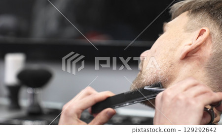 Professional barber grooming client's beard with precision, using comb and razor within stylish contemporary barbershop setting 129292642
