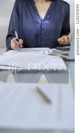 Businesswoman wearing blue dress is working with laptop, calculator and documents in a professional office setting. Low lighting, close-up vertical view. Business people, audit concept 129292649