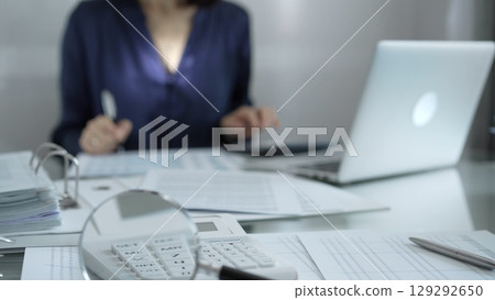 Magnifying glass, white calculator, pen and documents are lying on accountant's desk. Businesswoman wearing blue dress is working with documents on the background. Business and audit concept 129292650