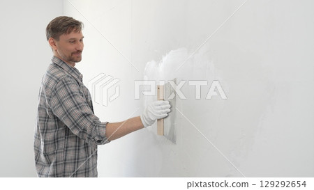 Man construction worker wearing protective gloves and plaid shirt, spreading plaster smoothly across wall surface using professional drywall taping knife during renovation project 129292654
